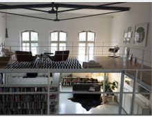 Zweistöckiger, moderner Wohnraum mit einer Galerieebene. Oben befinden sich Sitzmöbel auf einem schwarz-weiß gemusterten Teppich vor großen Rundbogenfenstern, unten ein Wohnzimmer mit Regalen, einer Couch, Pflanzen und modernen Beleuchtungselementen.