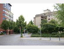 Straßenecke mit Kopfsteinpflaster, umgeben von mehrstöckigen Wohngebäuden; links ein modernes Gebäude mit roten und weißen Fassadenelementen, rechts ein beiges Gebäude mit Balkonen und umliegenden Bäumen. Verkehrszeichen und ein Straßenschild sind am Gehweg sichtbar.