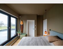 Helles Schlafzimmer mit großen bodentiefen Fenstern, die den Blick auf eine grüne Landschaft freigeben. An einer olivgrünen Wand hängt ein dekoratives Surfbrett; daneben sind eine Zimmerpflanze, weiße Türen und ein graues Bett mit beigen Kissen zu sehen.