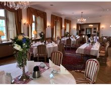 Eleganter Veranstaltungsraum mit gedeckten runden und rechteckigen Tischen, dekoriert mit Blumenarrangements und weißen Tischdecken. Der Raum verfügt über große Fenster mit Vorhängen, Kronleuchter an der Decke und eine Einrichtung im klassischen Stil.