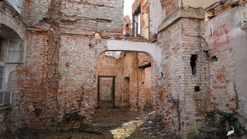 Innenhof einer verfallenen Backsteinruine mit freiliegenden, teils verputzten Mauern und einem gemauerten Rundbogen, der in einen weiteren Raum führt. Der Boden ist mit Moos und Schutt bedeckt, und mehrere Tür- und Fensteröffnungen sind ohne Rahmen oder Glas erhalten.