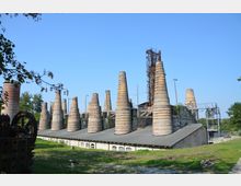 Historische Industrieanlage mit mehreren kegelförmigen, gemauerten Schornsteinen auf einem langen Dach. Im Hintergrund ist eine Metallkonstruktion sowie umliegendes Grünland erkennbar.