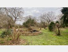 Ein weitläufiger Garten mit einer Rasenfläche, Bäumen ohne Laub und Sträuchern. Im Vordergrund steht ein rundes, gemauertes Beet mit einer Schubkarre darauf, rechts ein hölzernes Vogelhaus auf einem Pfahl.