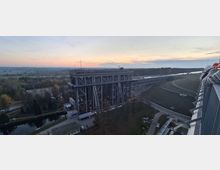 Luftaufnahme eines Schiffshebewerks in einer ländlichen Umgebung bei Sonnenuntergang. Zu sehen sind die massive, stahlverstärkte Konstruktion des Hebewerks, umliegende Wege, Bäume und ein angrenzendes Gewässer im Hintergrund.