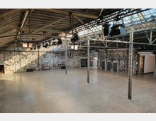 Geräumige, industrielle Halle mit Betonboden, blanken Ziegelwänden und einem Dach aus Metallträgern sowie großen Oberlichtern, die das Innere mit Tageslicht erhellen. An der Decke befindet sich eine Beleuchtungsanlage; mehrere Türen und ein Seiteneingang sind sichtbar.