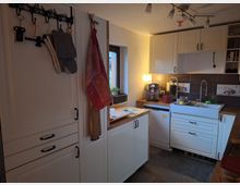 Kleine Küche mit weißen Schränken und Holzarbeitsflächen. Ein Waschbecken, eine Kaffeemaschine und eine Spülmaschine sind sichtbar; an der Wand hängen Küchenutensilien und Handtücher.