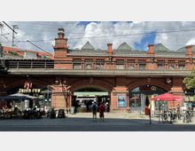 Backsteinbahnhof mit bogengewölbten Eingängen, dem Schriftzug „Hackescher Markt“ und angrenzenden Geschäften, darunter ein Irish Pub und eine Bäckerei. Im Vordergrund stehen Tische mit Sitzenden, Fahrräder und zwei Menschen vor dem Haupteingang.