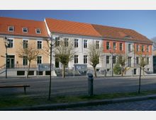 Straßenansicht eines Gebäudes mit drei unterschiedlich gefärbten Fassaden in Gelb, Weiß und Rot, jeweils mit roten Ziegeldächern. Vor dem Gebäude führen Treppen zu den Eingängen, und es stehen mehrere kahle Bäume sowie traditionelle Straßenlaternen auf einem gepflasterten Platz.