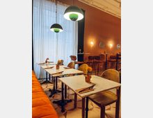 Ein gemütliches Café mit orangefarbener Wand, weißen Marmortischen und Vintage-Holzstühlen. Hängende runde Lampen beleuchten den Raum, und gelbe Blumen in kleinen Vasen dekorieren die Tische.