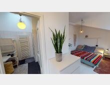 Ein heller Wohnraum mit Blick auf ein Schlafzimmer und ein Badezimmer. Im Vordergrund steht eine Zimmerpflanze auf einer weißen Kommode, das Schlafzimmer zeigt ein Doppelbett mit einer mehrfarbigen Decke, das Badezimmer verfügt über weiße Fliesen, eine Duschkabine und einen Handtuchwärmer.