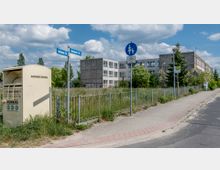 Auf einem Gehweg mit einer Bushaltestelle und einem Verkehrszeichen für Fußgänger und Radfahrer steht ein Kleider- und Schuhcontainer. Im Hintergrund sind ein Zaun, eine verwilderte Wiese sowie ein graues, mehrstöckiges Gebäude aus Betonplatten sichtbar.