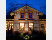 Zweistöckiges Gebäude im Stil eines historischen Backsteinbaus mit leuchtenden Fenstern, aufgenommen in der Dämmerung. Das Haus verfügt über Satteldach, detaillierte Fassadenelemente und ist von spärlicher Vegetation umgeben.