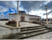 Ein zweistöckiges Gebäude mit heller Fassade und der Aufschrift "Bierstube" über dem Eingang, umgeben von Treppen, Geländern und gepflegten Sträuchern. Der Himmel ist bewölkt, und im Hintergrund sind Bäume und moderne Gebäude sichtbar.
