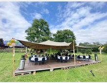 Offenes Festzelt mit sandfarbenem Sonnensegel auf einem Holzpodest, umgeben von einer grünen Wiese und Bäumen. Runde Tische mit blauen Stühlen und Dekorationen wie Luftballons und Lichterketten sind sichtbar.