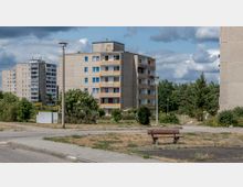 Wohngebiet mit Plattenbauten aus Beton in einer urbanen Umgebung, umgeben von Grünflächen und Bäumen. Im Vordergrund befindet sich eine kleine gepflasterte Fläche mit einer Holzbank, während im Hintergrund mehrere mehrstöckige Wohngebäude zu sehen sind.