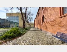 Gepflasterter Weg zwischen einem modernen Gebäude mit großen Glasfronten auf der linken Seite und einer roten Backsteinfassade auf der rechten Seite. Der Weg ist von Bäumen und Grünflächen gesäumt, im Hintergrund sind weitere gelbe Gebäude zu sehen.