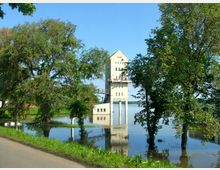 Ein modernes Gebäude auf hohen Säulen steht über einem Teich oder überfluteten Bereich, umgeben von grünen Bäumen und Wiese. Im Hintergrund ist ein klarer blauer Himmel zu sehen.
