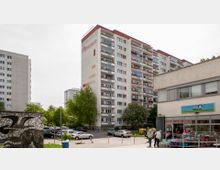 Hochhaussiedlung mit einem mehrstöckigen Wohngebäude im Zentrum, umgeben von Bäumen und parkenden Autos. Im Vordergrund befindet sich ein kleines Geschäft mit Schildern und Auslage, sowie vereinzelte Passanten und eine gemusterte Mauer links im Bild.