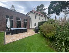 Wohnhaus mit zwei unterschiedlichen Fassaden: eine moderne Backsteinwand mit großen Glasfenstern und eine helle verputzte Hausseite mit roten Dachziegeln. Im Vordergrund eine gepflasterte Terrasse mit Gartenmöbeln und Sonnenschirm, umgeben von gepflegtem Rasen, Büschen und hohen Gräsern.