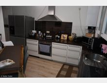 Moderne Küche mit weißen Unterschränken, dunkler Arbeitsplatte und Edelstahlgeräten, darunter ein großer Kühlschrank und ein Dunstabzug über einem Kochfeld. Rechts ist ein runder Spülbereich, während links ein Esstisch mit Sitzgelegenheiten zu sehen ist.