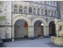 Blick auf einen historischen Arkadengang mit Rundbögen und einer Fassade aus hellem Stein, darüber Reihenfenster mit Bögen. Im Vordergrund steht ein steinerner Brunnen mit Löwenskulptur.