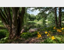 Ein grüner Park mit einem Teich, umgeben von Bäumen und Sträuchern. Im Vordergrund stehen gelbe Blumen, große Steine sowie ein Baumstamm, während Enten auf den Steinen am Wasser sitzen.