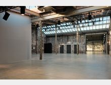 Industriehalle mit modernisiertem Innenraum, geprägt von Betonboden, freiliegenden Stahlträgern und großen Glasdachfenstern, die viel Tageslicht einlassen. Wände aus unverputztem Ziegelstein und mehrere dunkle Türen sowie Beleuchtungstechnik an der Decke weisen auf eine Nutzung als Veranstaltungsort hin.