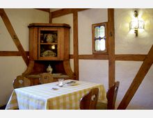 Rustikaler Raum mit Fachwerkstruktur, einer kleinen Vitrine aus Holz mit Keramikgeschirr und einem Tisch mit gelb-weiß karierter Tischdecke. An der Wand ein kleines Fenster mit farbigen Akzenten und eine beleuchtete Wandleuchte.