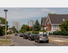 Wohnstraße mit geparkten Autos am Straßenrand, flankiert von Einfamilienhäusern mit Gärten auf der rechten Seite und Bäumen auf der linken Seite; im Hintergrund sind mehrstöckige Wohngebäude zu sehen. Die Szene wird von einem Straßenschild mit der Aufschrift "Amanlisweg" und einzelnen Straßenlaternen ergänzt.