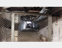 Ein mit Maschendraht verschlossenes Untergeschoss oder Kellerbereich, durch den Rohre und Leitungen an der Decke verlaufen; im Hintergrund ist ein schwach beleuchteter Bereich sichtbar.