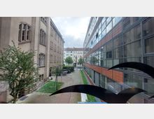 Blick auf einen schmalen Innenhof mit zwei Gebäuden: links ein Altbau mit hellbeiger Fassade und großen Fenstern, rechts ein moderner Bau mit einer Fassade aus Glas und orangefarbenen Paneelen. Im Hintergrund sind weitere Wohngebäude sowie Bäume und ein gepflasterter Weg sichtbar.