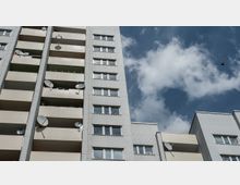 Plattenbau-Wohngebäude mit mehreren Balkonen, an denen Satellitenschüsseln angebracht sind, vor einem Himmel mit einigen Wolken und einem fliegenden Vogel. Die Fassade besteht aus hellen Paneelen mit rechteckigen Fenstern.