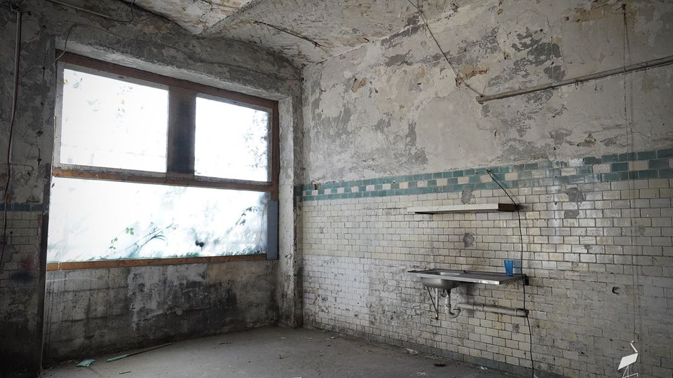 Verlassener, stark beschädigter Innenraum mit abblätterndem Putz, gemauerter Tonnenwölbung und großem, milchigem Fenster an der linken Wand. An der rechten Wand befinden sich weiße Wandfliesen mit blaugrünem Streifen sowie ein kleines Waschbecken mit Ablage.