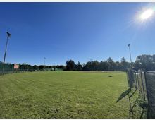 Weitläufiges Sportfeld mit gepflegtem grünem Rasen, umgeben von Zäunen und umstehenden Bäumen im Hintergrund. Vereinzelte Flutlichtmasten ragen in den blauen, sonnigen Himmel.