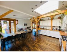 Offene Wohnküche mit Holzdetails und großen Fenstern. Weiße Küchenzeile mit Holzarbeitsplatten, dunkler Holzboden, ein Esstisch mit schwarzen Stühlen, und eine Sitzecke mit Sofas; dekoriert mit hängenden Pflanzen und natürlichem Licht durch ein Dachfenster.