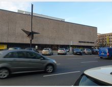 Außenansicht der Deutschen Oper Berlin, ein moderner, rechteckiger Bau mit grobkörniger Fassade. Vor dem Gebäude befinden sich parkende Autos, eine viel befahrene Straße und eine abstrakte Metallskulptur.