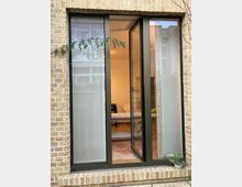 Fensterfront mit dunkelbraunen Rahmen in einer Fassade aus gelben Ziegelsteinen, teilweise geöffnet und mit Blick in einen Innenraum mit heller Beleuchtung. Ein kleiner Blumentopf und Kletterpflanzen befinden sich am Rand des Fensters.