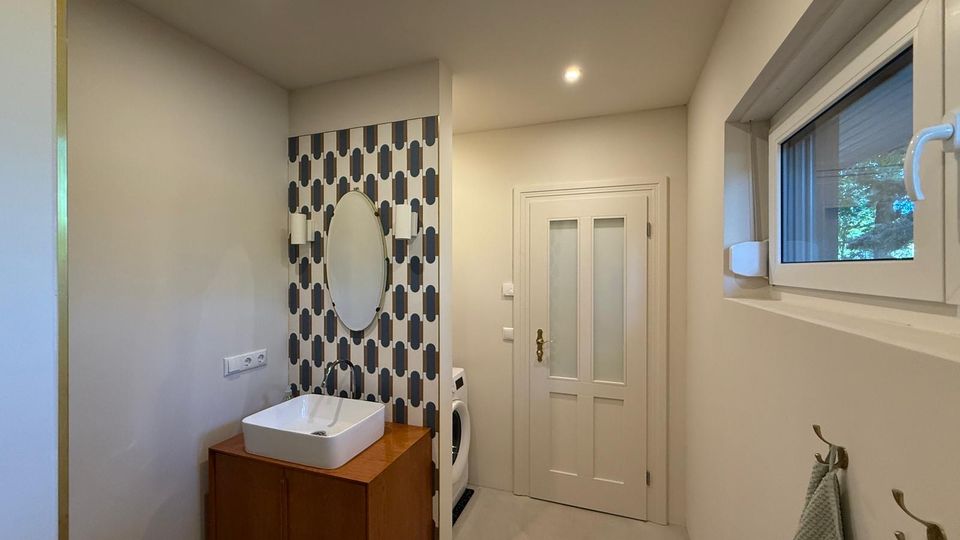 Modernes Badezimmer mit hellem Design, einer Waschbeckeneinheit aus Holz, ovalem Spiegel und gemusterter Fliesenrückwand. Rechts an der Wand befindet sich ein Fenster und darunter Handtuchhaken, im Hintergrund eine weiße Tür und eine Waschmaschine.