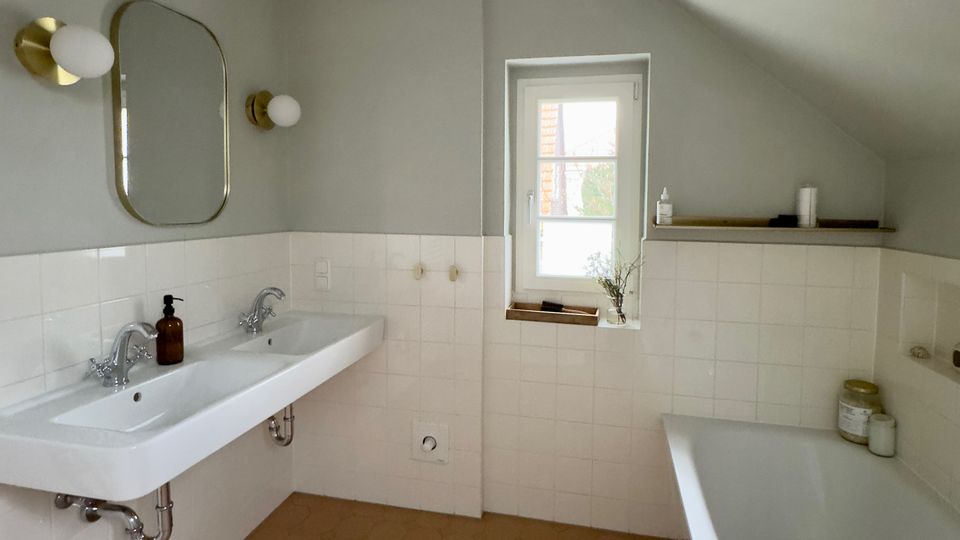 Helles Badezimmer mit schräger Dachdecke, weißen Wandfliesen und beigefarbenem Fliesenboden mit sechseckigem Muster. Links ein Doppelwaschbecken mit Spiegel und Wandleuchten, rechts eine Badewanne; mittig ein kleines Fenster über einer schmalen Ablage.