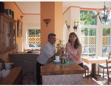 Ein kleines Café oder Restaurant mit großen Fenstern, die viel Tageslicht hereinlassen. Im Vordergrund stehen eine Frau und ein Mann an einer Theke aus Stein, im Hintergrund sind Tische und Stühle sichtbar.