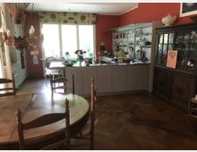 Offene Wohnküche mit einem großen runden Holztisch und rustikalen Stühlen im Vordergrund, einer Kücheninsel mit modernen grauen Schränken in der Mitte und einem antiken Holzschrank rechts. Der Raum hat rote Wände, ein großes Fenster mit Tageslicht und Parkettboden.