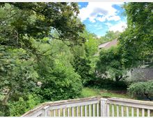 Ein grüner Garten mit dichtem Laub von Bäumen und Sträuchern, im Hintergrund Dächer von Wohnhäusern mit roten Ziegeln. Im Vordergrund ist ein verwittertes Holzgeländer sichtbar.