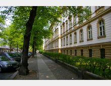 Straßenansicht mit einem historischen Gebäude im Stil der klassischen Architektur, das durch weiße Fassade und verzierte Fensterrahmen gekennzeichnet ist, links gesäumt von Bäumen und geparkten Autos. Ein Gehweg verläuft parallel zum Gebäude, flankiert von niedrigen Hecken.