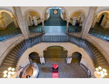 Innenansicht eines historischen Gebäudes mit einem runden, mehrfach geteilten Treppenaufgang aus Stein, dekorativen Bögen und schmiedeeisernen Geländern. Im Zentrum des gefliesten Bodens steht ein rotes Herz auf einem Podest, flankiert von zwei Info-Ständen; im Hintergrund befindet sich eine Uhr über einer breiten Treppe.