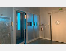 Moderner Innenraum mit einer mattierten Glastür und Wandpaneelen, durch die blaues Licht dringt. Links eine Tür mit metallischer Oberfläche und rechts eine Schiebetür mit Symbol. An der Wand befinden sich Gegensprechanlagen, ein Regal mit Gegenständen, ein Feuerlöscher und ein Desinfektionsmittelspender.