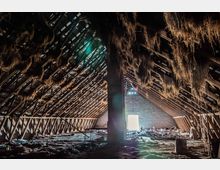 Innenansicht eines großen, verlassenen Dachbodens mit freiliegender Holzkonstruktion. Trockenes Stroh hängt von den Balken herab, und der Boden ist mit Schutt sowie verschiedenen Gegenständen bedeckt; im Hintergrund ist ein heller, rechteckiger Ausgang sichtbar.