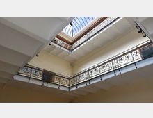 Blick von unten auf einen offenen Innenhof eines Gebäudes mit zwei Etagen. Die Etagen sind von kunstvoll verzierten schmiedeeisernen Geländern umgeben, und die Decke schließt mit einem transparenten Glasdach ab, das Licht hereinlässt.