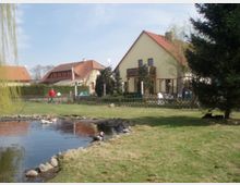 Ein kleines Grundstück mit einem Teich im Vordergrund, in dem Enten schwimmen. Im Hintergrund stehen zwei Wohnhäuser mit roten Ziegeldächern, umgeben von Bäumen, einer Hecke und einem Zaun.