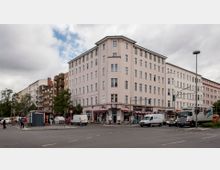 Straßenkreuzung mit einem fünfstöckigen Wohn- und Geschäftshaus im Fokus, das eine helle Fassade und zahlreiche Fenster hat. Im Erdgeschoss des Gebäudes befinden sich Läden und Cafés, vor denen geparkte Autos, Lieferwagen und Fußgänger zu sehen sind, während an der Kreuzung Ampeln und ein kleines Toilettenhäuschen stehen.