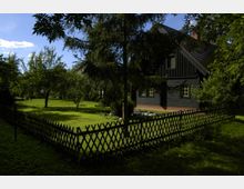 Dunkler Holzbauernhof mit grünen Fensterrahmen, umgeben von einem Garten mit Rasen, Bäumen und einem Holzzaun. Der Bereich ist schattig, mit sonnigen Stellen, und der Himmel ist klar.